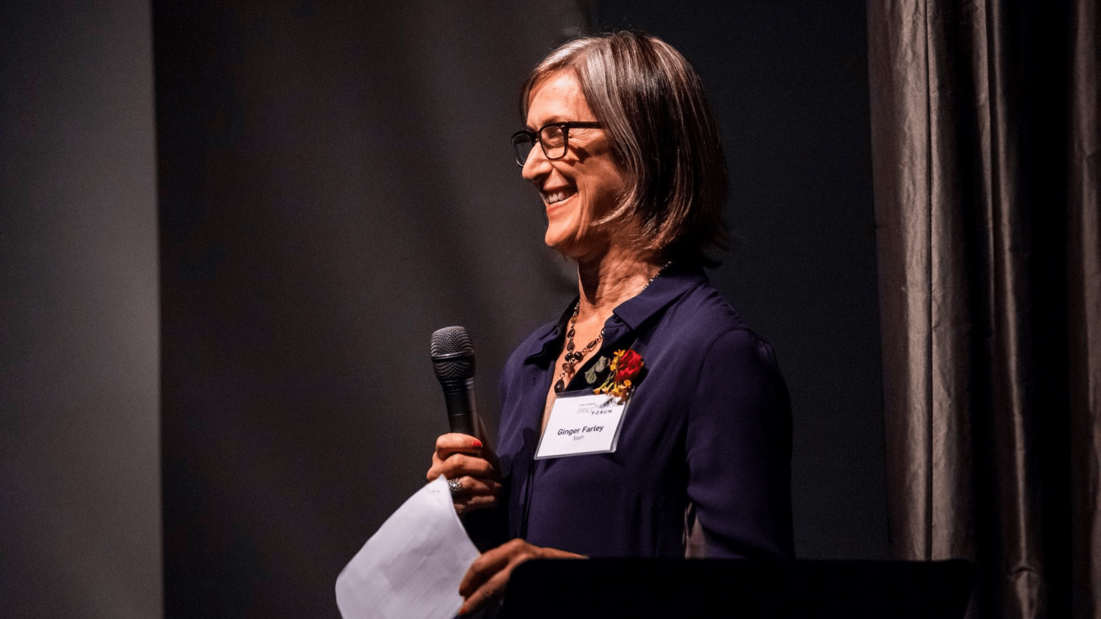 A woman with a navy blouse, flower boutonniere, and glasses smiles holding a microphone and sheet of paper