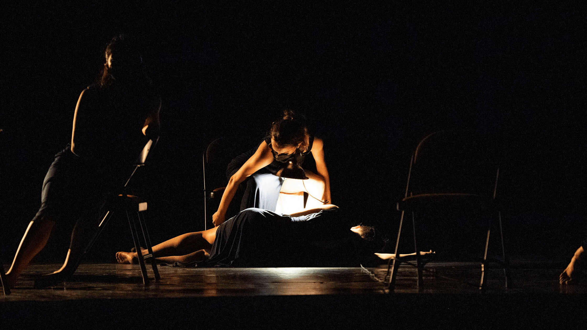 Four dancers on a darkly lit stage. Two of them are in chairs. One is laying on her side facing upstage. One is more visible, with a hand-held light.