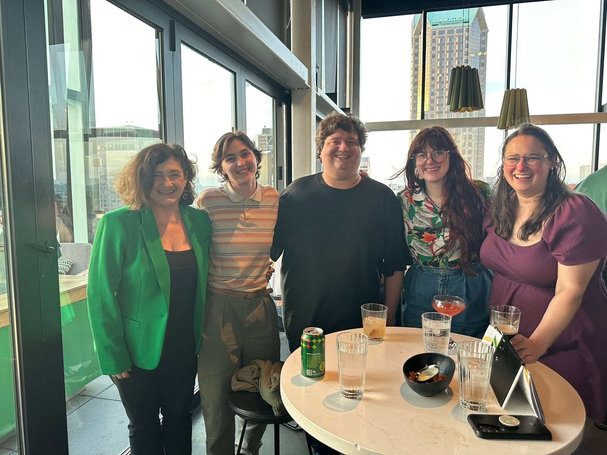 Five Irish singers pose together at a rooftop social event with drinks and appetizers on a high-top table. Large windows overlook the downtown skyline as evening sunlight streams into the venue.