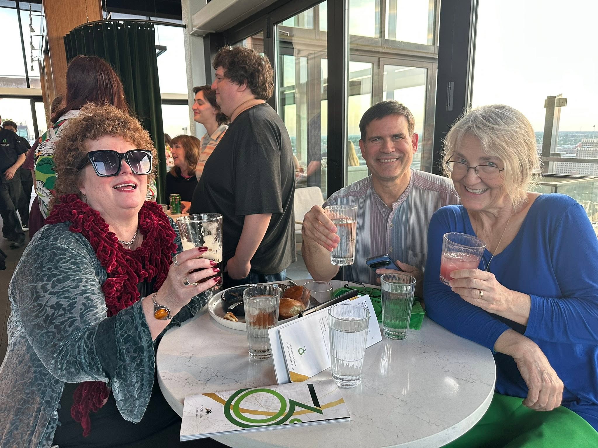 Three Irish singers enjoy drinks and conversation at a rooftop social event, smiling toward the camera while seated at a high-top table. Large windows provide views of the downtown skyline, with other guests socializing behind them.