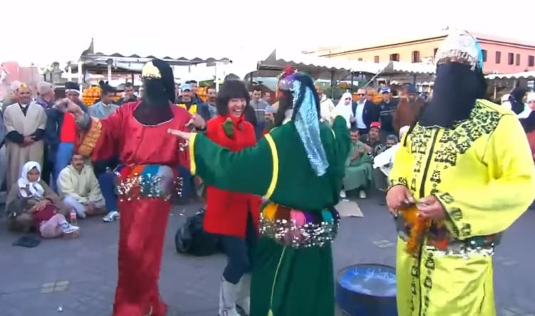 一個傳統的摩洛哥表演場景,人們在室外圍觀兩位穿著鮮艷服裝的舞者,包括一位身著紅色服裝的舞者和一位身著綠色服裝的舞者,他們正在表演舞蹈,周圍有觀眾聚集。