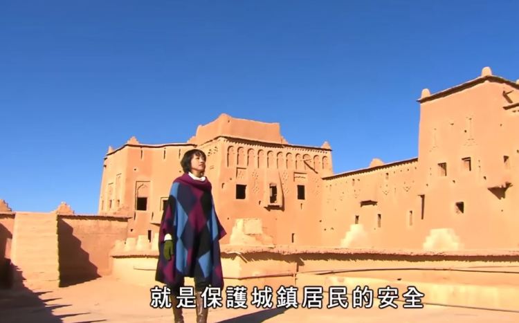 A woman standing in front of a traditional adobe building in a desert setting, wearing a colorful poncho, with a clear blue sky in the background.