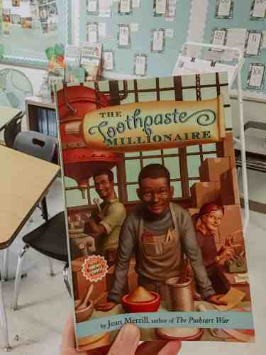 A teacher holding up the novel "Toothpaste Millionaire" by Jean Merrill.