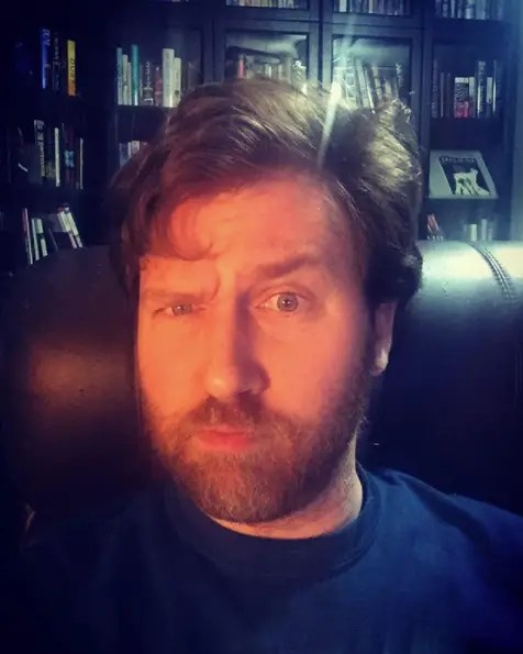 Photo of Christopher Opyr withone eyebrow raised, sitting in front of a bookshelf backdrop.