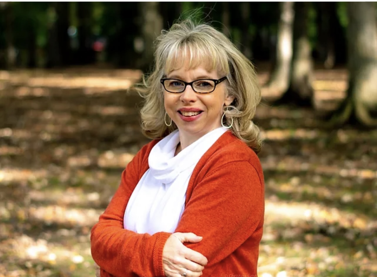 Sharon Waldrop outdoors among autumn trees, arms folded, smiling at the camera in a rust-colored cardigan.