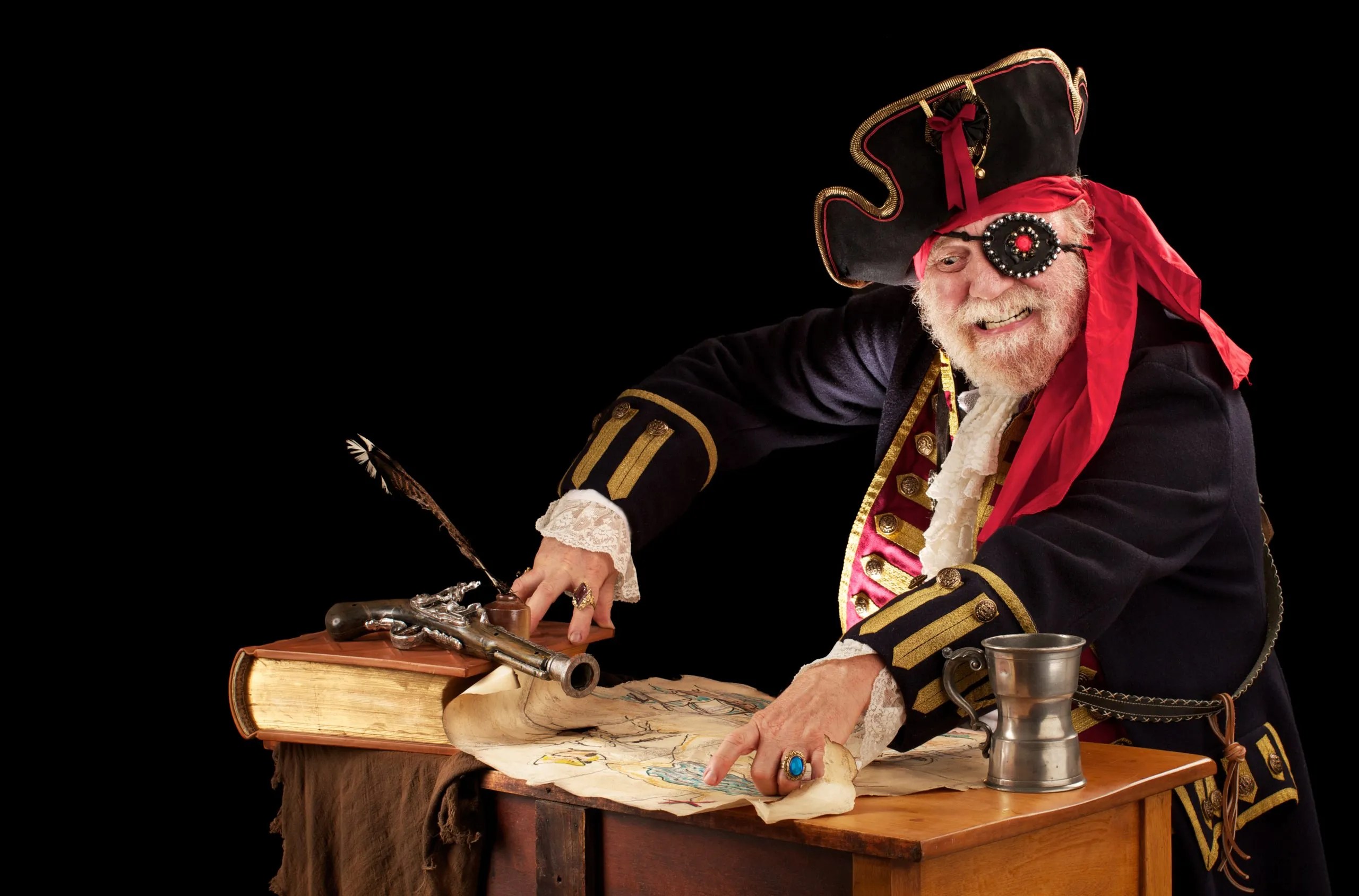 Gleeful old pirate captain sits at his table with still life of log book, quill pen, musket, pewter mug, and tattered weather beaten treasure map. He is wearing an authentic looking 19th century pirate costume with jewelled eye patch, braided topcoat perusing his treasure map.