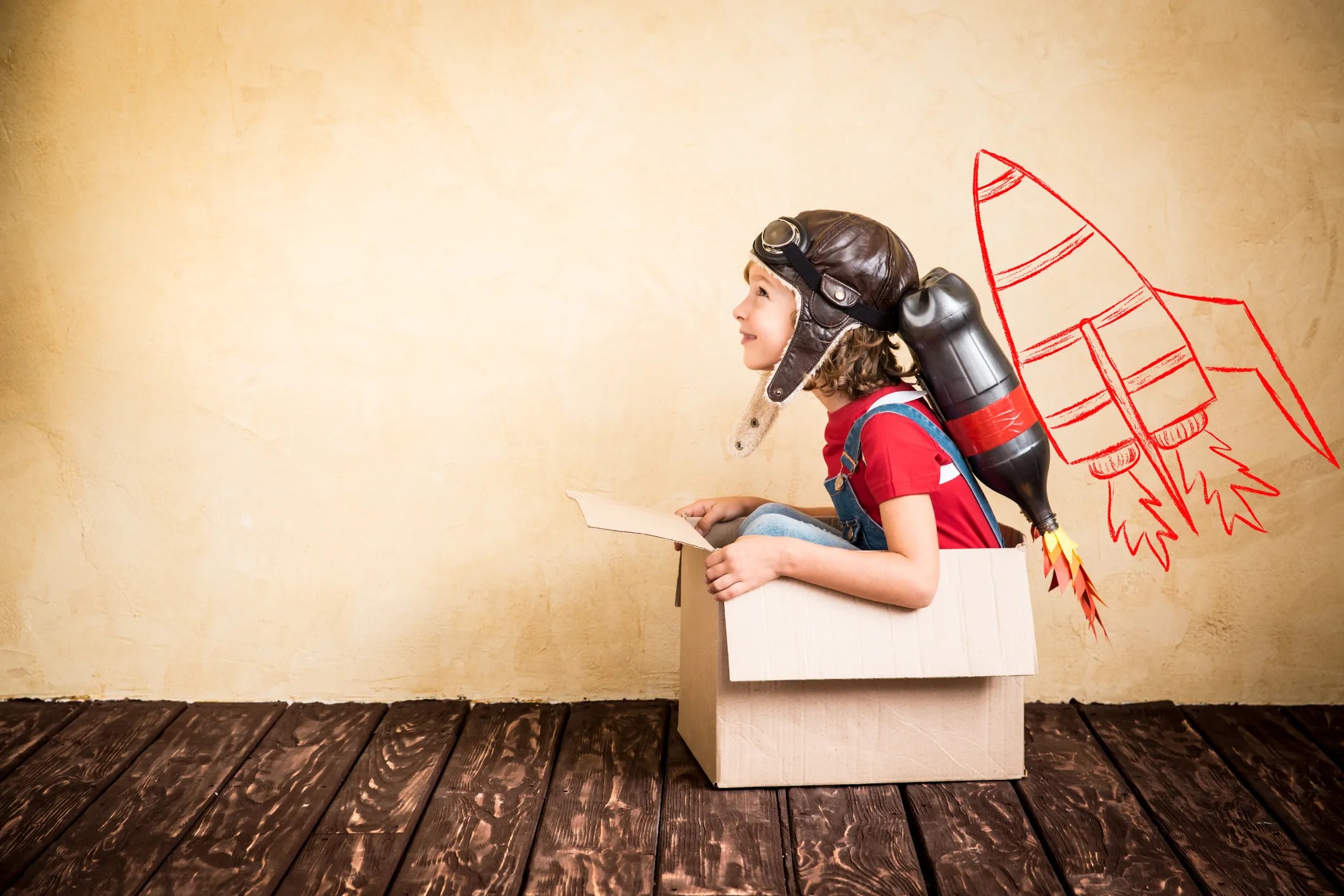 A young kid in a card board box fantasying he has a rocket strapped to his back.
