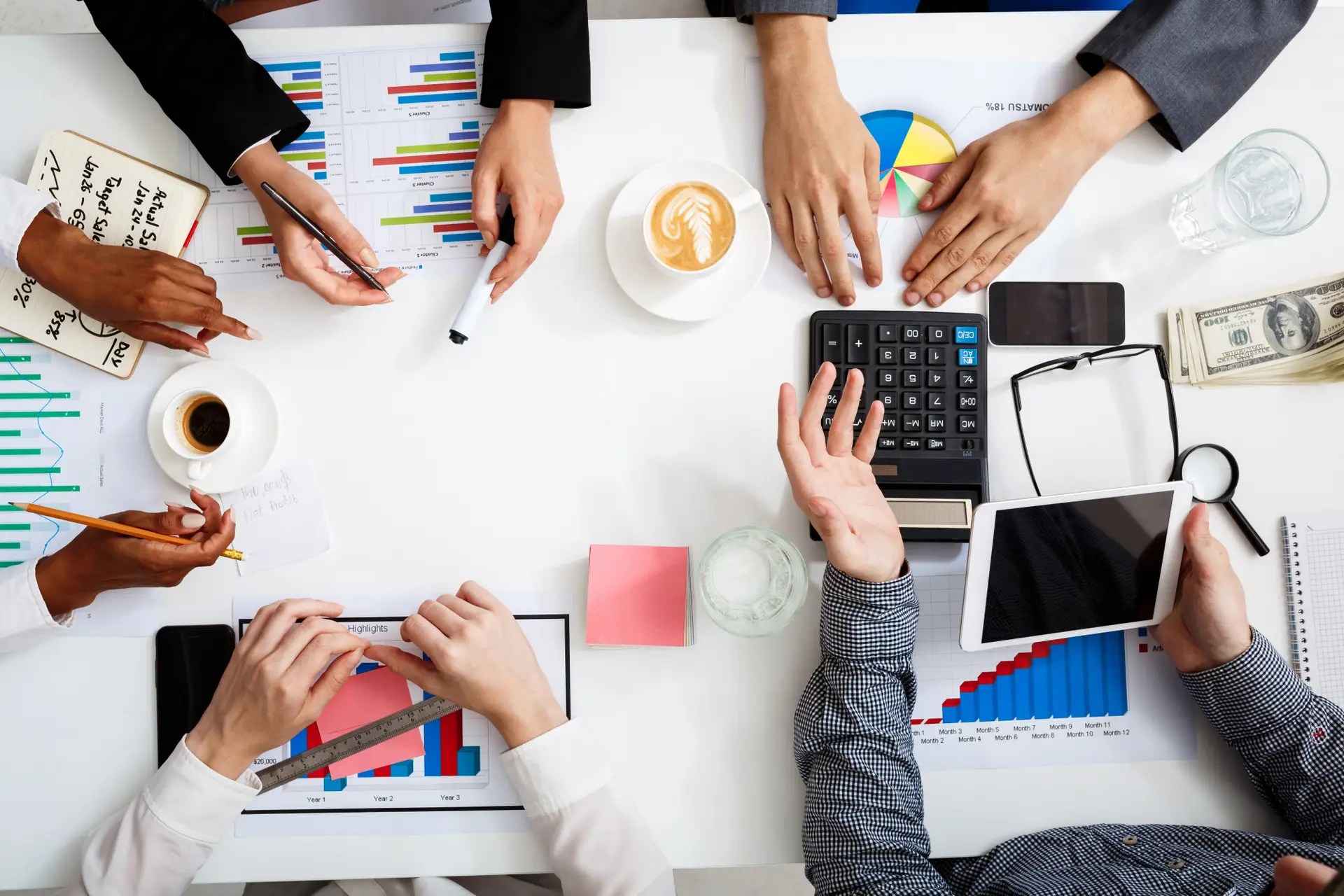 Overhead view of a business meeting with several hands around a table, reviewing colorful charts and graphs, plus coffee and devices on the desk.