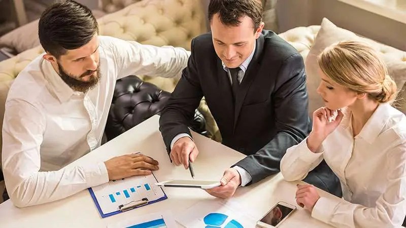 Three professionals in a meeting discussing charts on a white table with a tablet and a smartphone