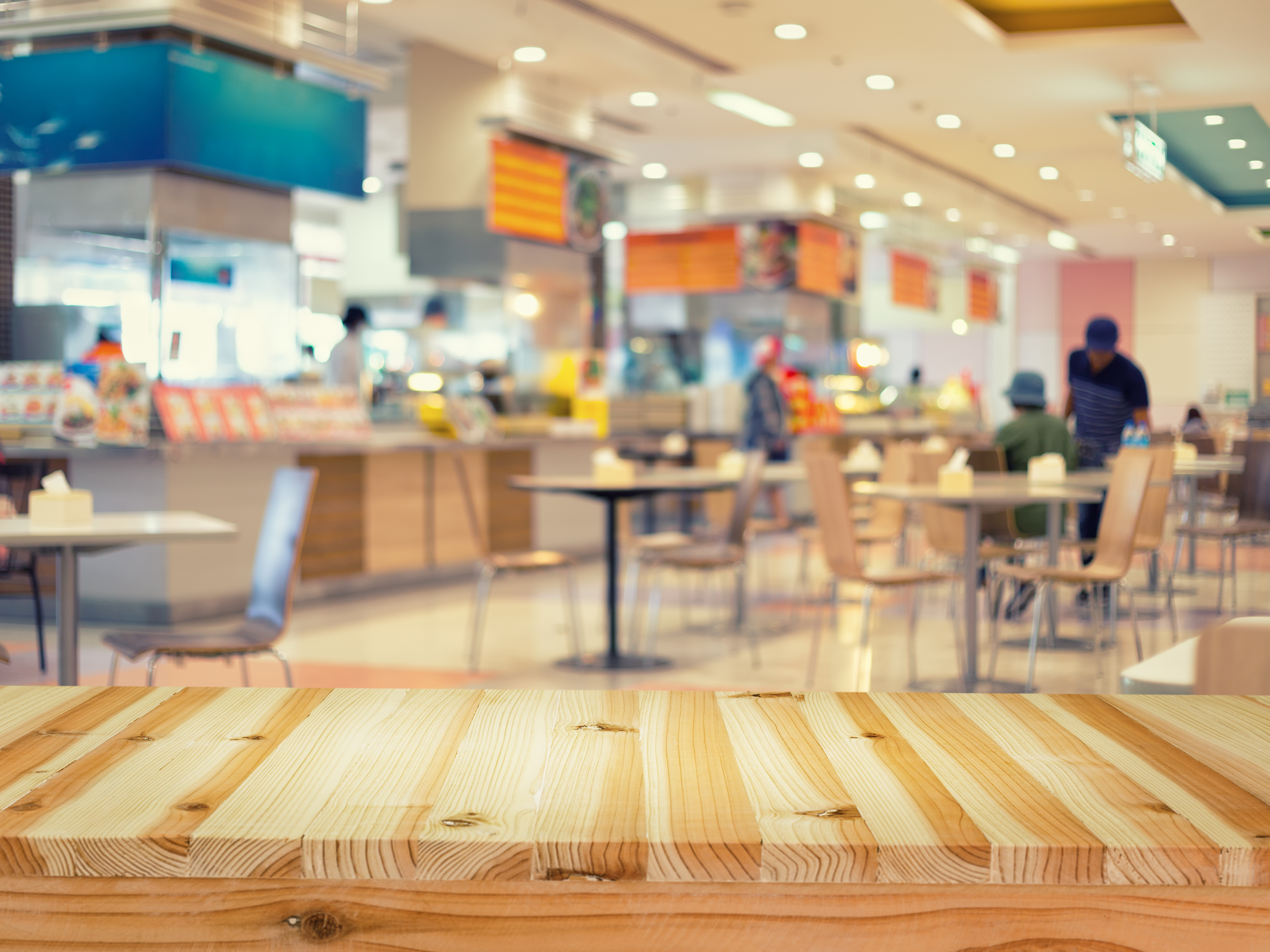 A blurred view of a brightly lit restaurant or food court with tables, chairs, and customers in the background, and a wooden tabletop in the foreground.