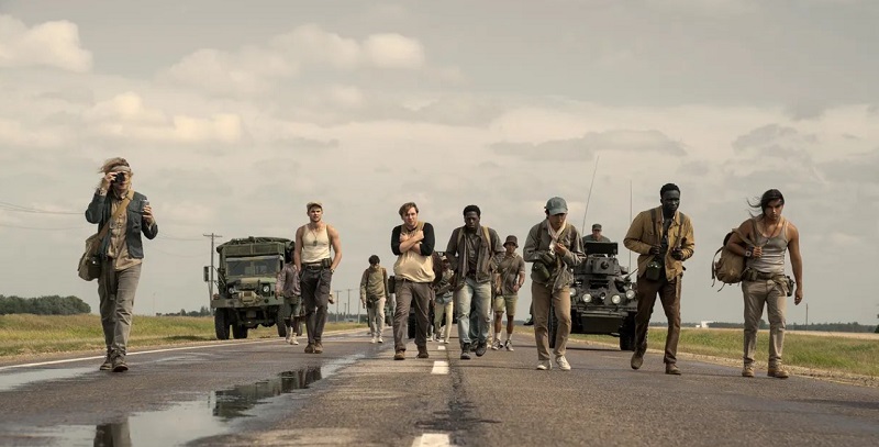 Still from The Long Walk movie. Hoffman, Jonsson and others walk as a group down a road. Military vehicles flanking them on either side.