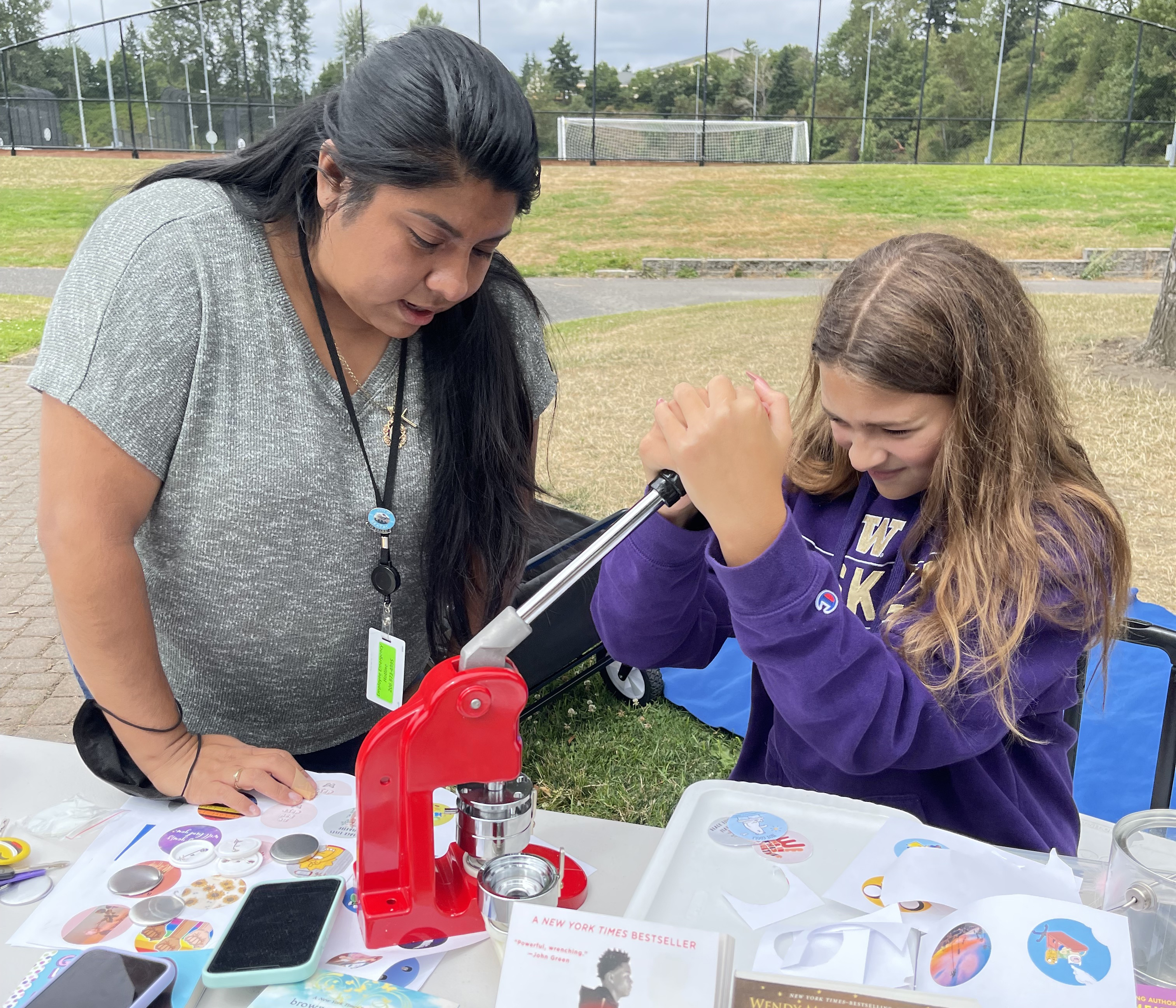 A girl presses down on a lever for a button maker as a woman watches.