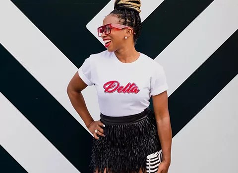 African American woman smiling in front of a black and white diamond wall, wearing a white short-sleeve t-shirt with the word "Delta" on the front. T-shirt by JLS Presents (jlspresents.com).