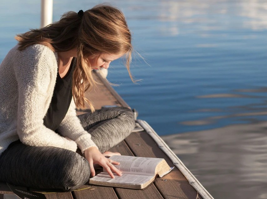 A girl sitting by the water reading her Bible to avoid stress