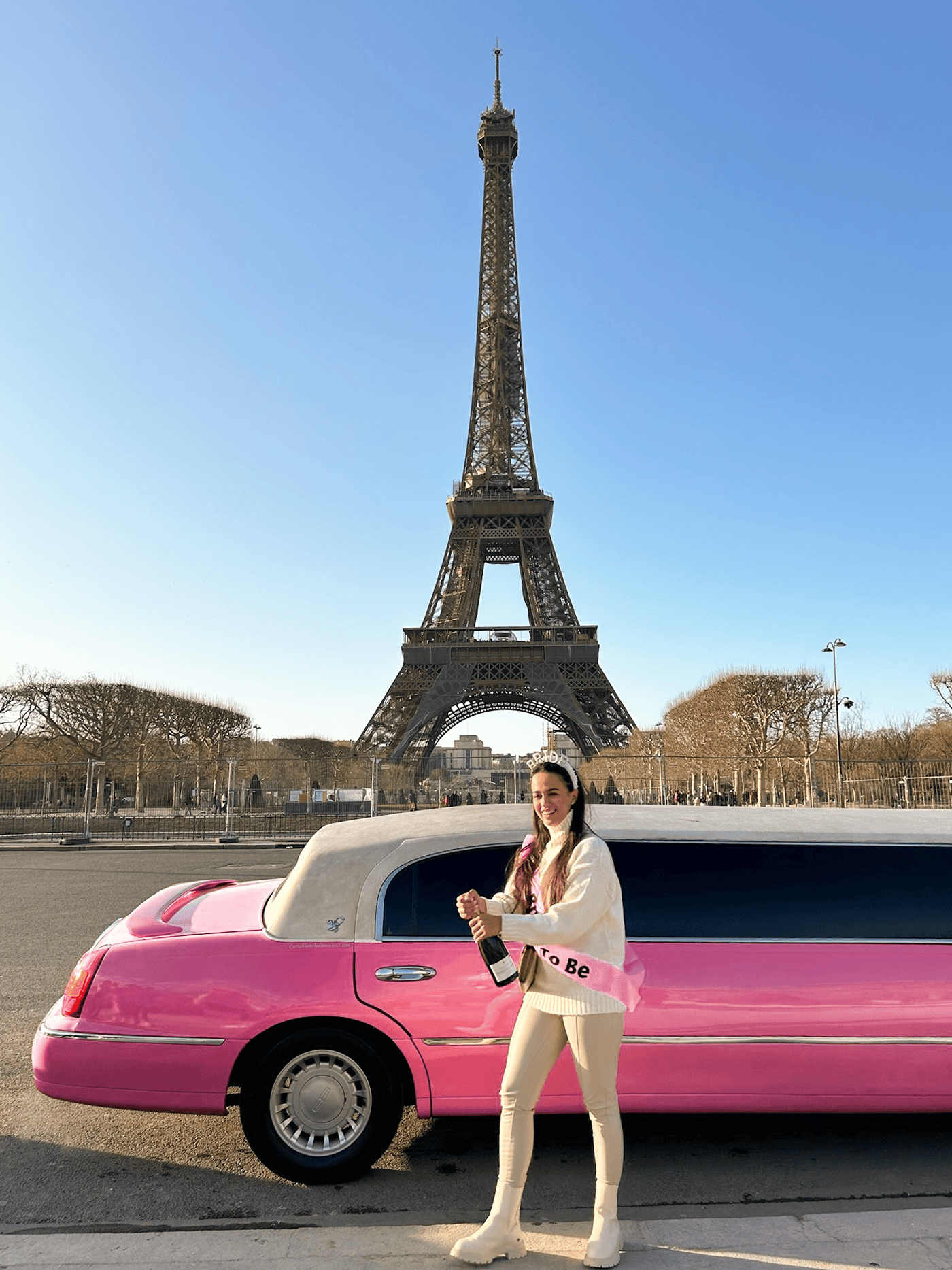 Girl standing in front of a pink limo with a bottle of champagne. In the background, the Eiffel tower and a blue sky.