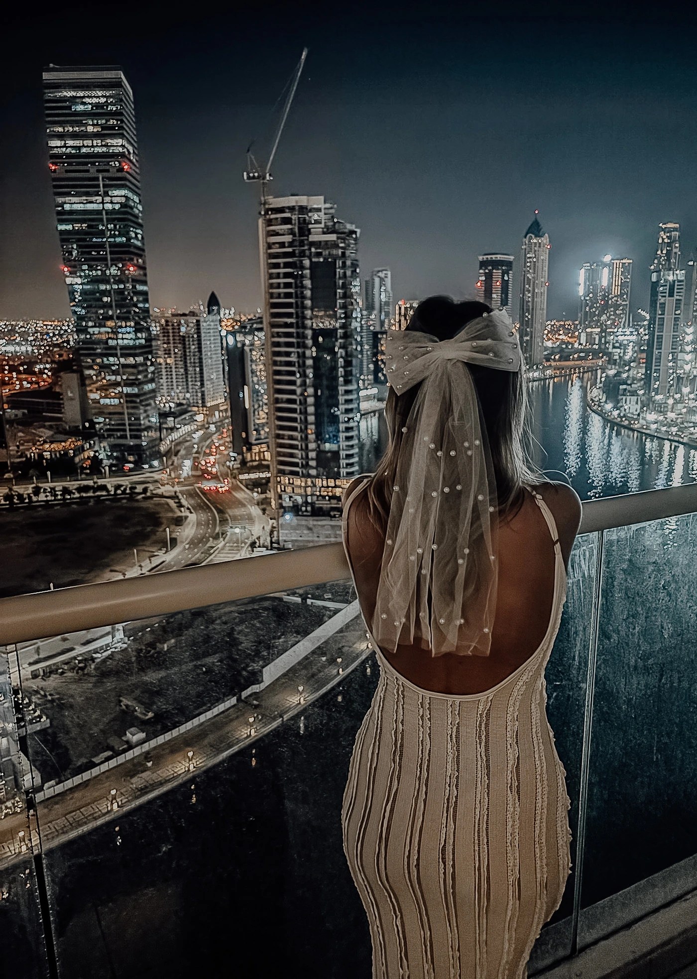A girl admiring the city skyline view at night on her Dubai hen do, wearing a white dress and bow veil
