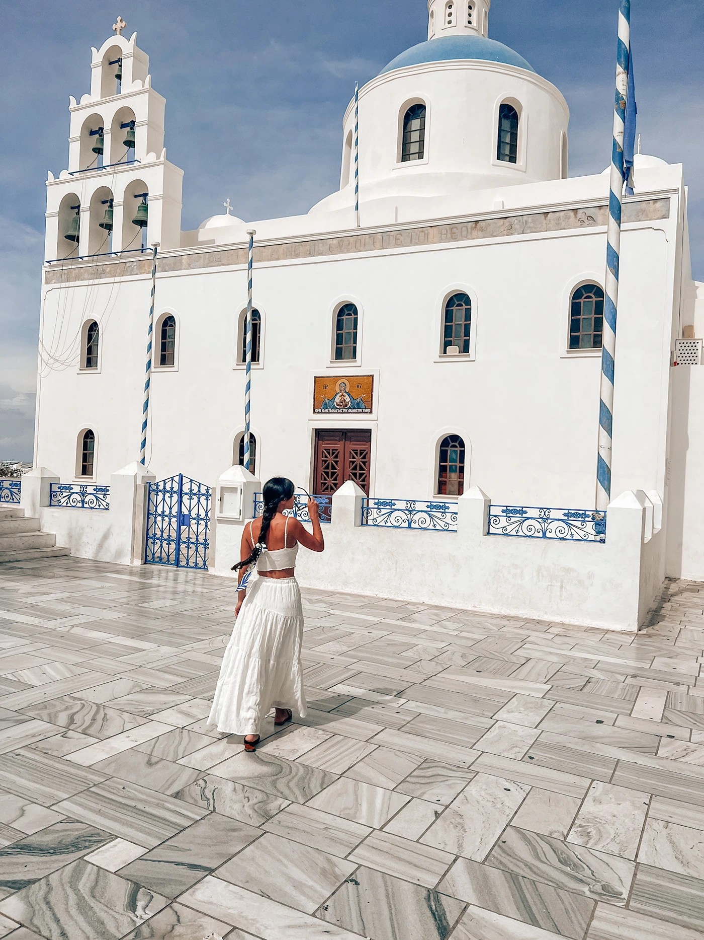 A girl in a long white skirt and top walking towards a church with a blue dome and bells in Santorini in September
