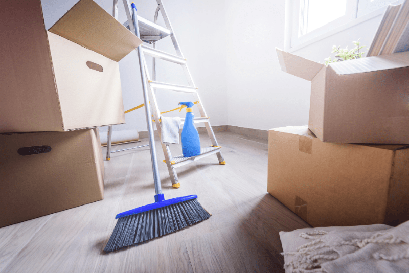 packing boxes shown in white walled room with wooden floor, next to ladder, broom and cleaning spray