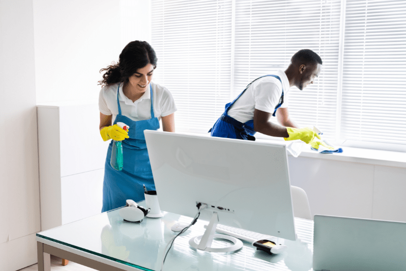 a male cleaner and a female cleaner, both wearing rubber gloves, cleaning an office desk and a windowsill using cloths and cleaning spray