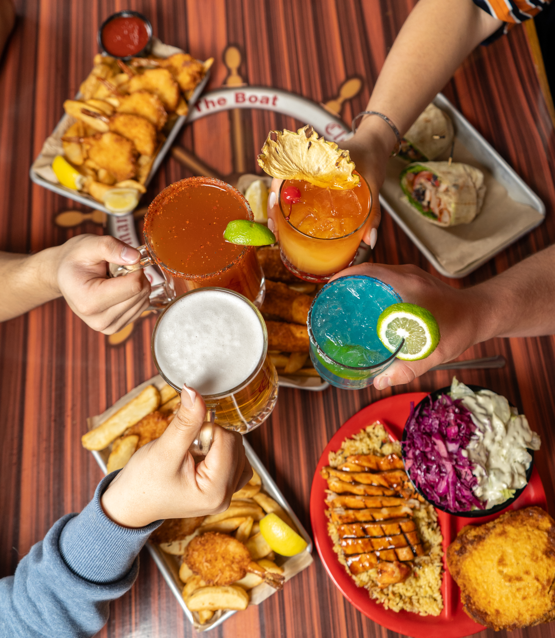 Friends clink a variety of beers and cocktails over a table covered in Galley dishes like coconut shrimp and wraps.