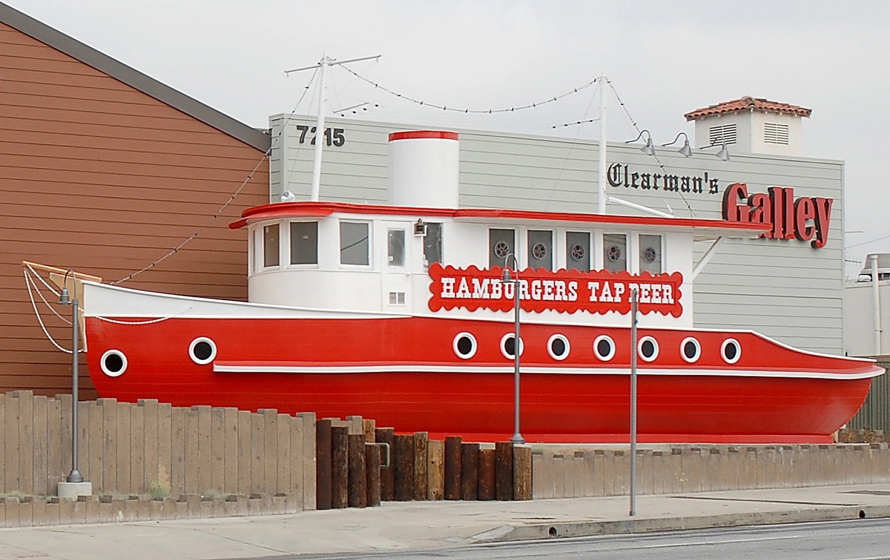 A vibrant red boat named 'The Boat' is displayed on a sidewalk, featuring rounded windows and a rounded roof. The boat is situated next to a wooden fence and surrounded by small plants, while a light-colored building is visible in the background.