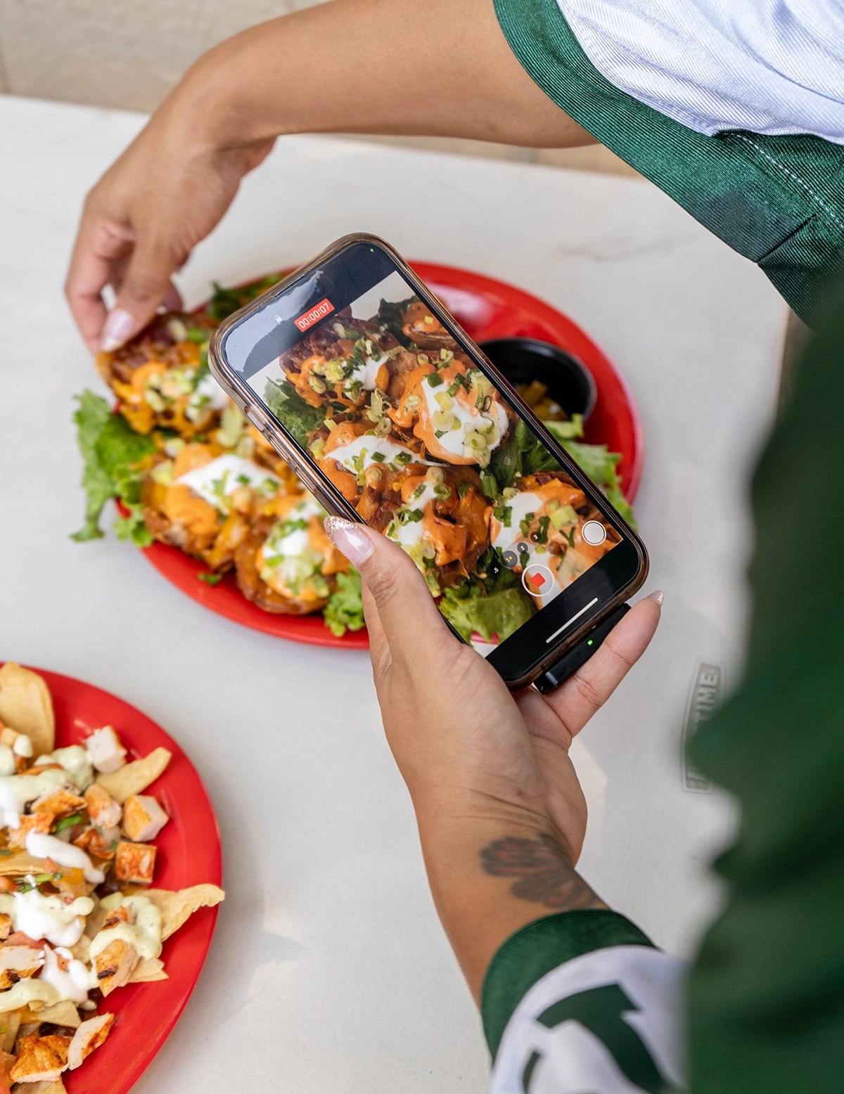 A person is taking a photo of a plate of potato skins topped with cheese, sour cream, and green onions, while another plate of nachos is visible in the foreground.