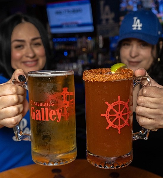 Two women are holding up large mugs of drinks at a table. One mug has a beer, while the other is a michelada with a rim. Both mugs have the logo "Clearman's Galley."