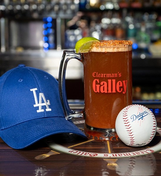 A blue LA Dodgers cap, a large glass of michelada with a lime wedge, and a baseball with "Dodgers" written on it, set against a blurred bar background.