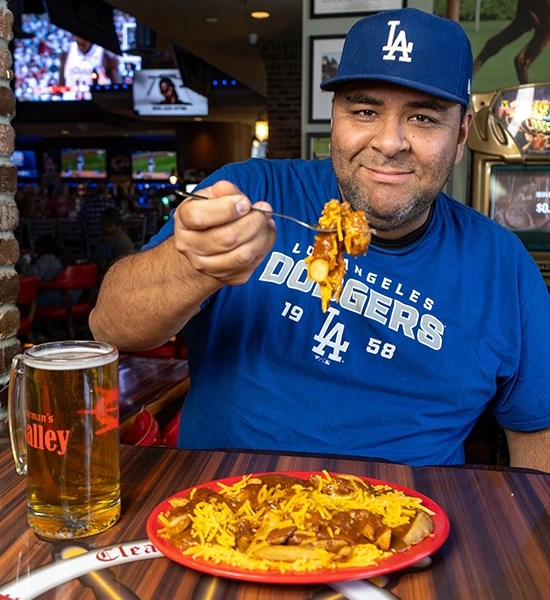 A man holding a fork with food in a casual dining setting, wearing a blue Los Angeles Dodgers shirt and a matching cap, with a pint of beer and a plate of chili cheese fries in front of him.