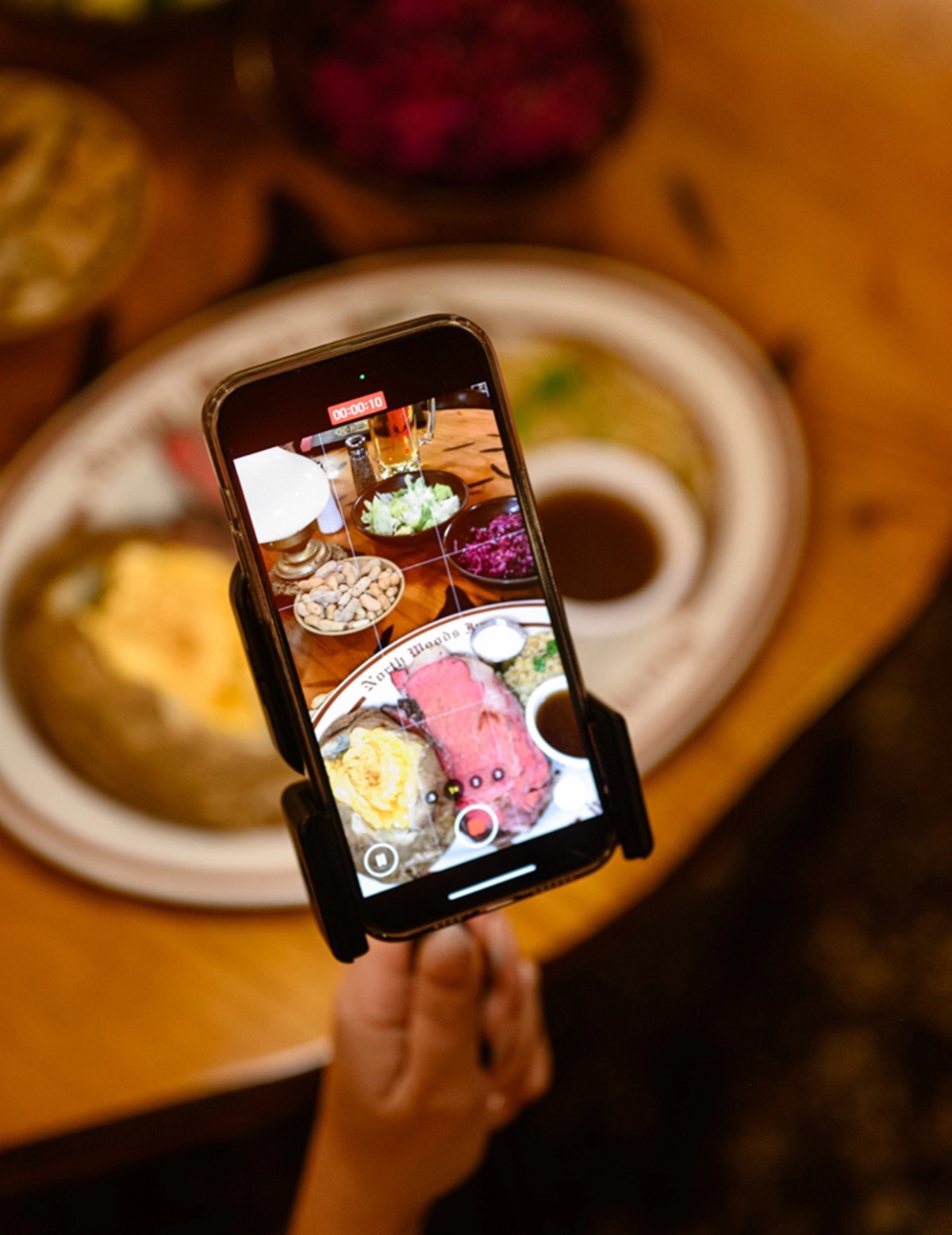 A hand holding a smartphone is capturing a video of a plate of food, which includes colorful dishes, salad, and sauces, on a wooden table.
