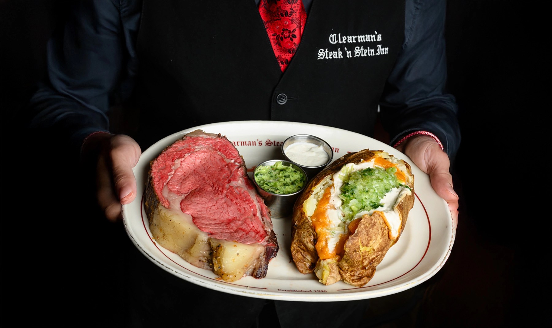 A plate with a large cut of prime rib, a loaded baked potato topped with sour cream and green onions, and two small cups of sauce, held by a person wearing a black vest.