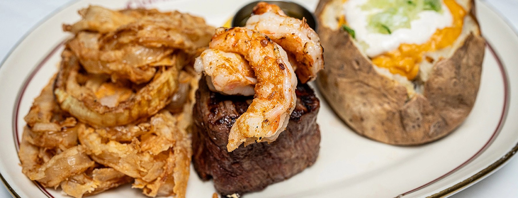 A plate featuring grilled shrimp on a beef steak, accompanied by crispy onion rings and a baked potato topped with cheese and sour cream.