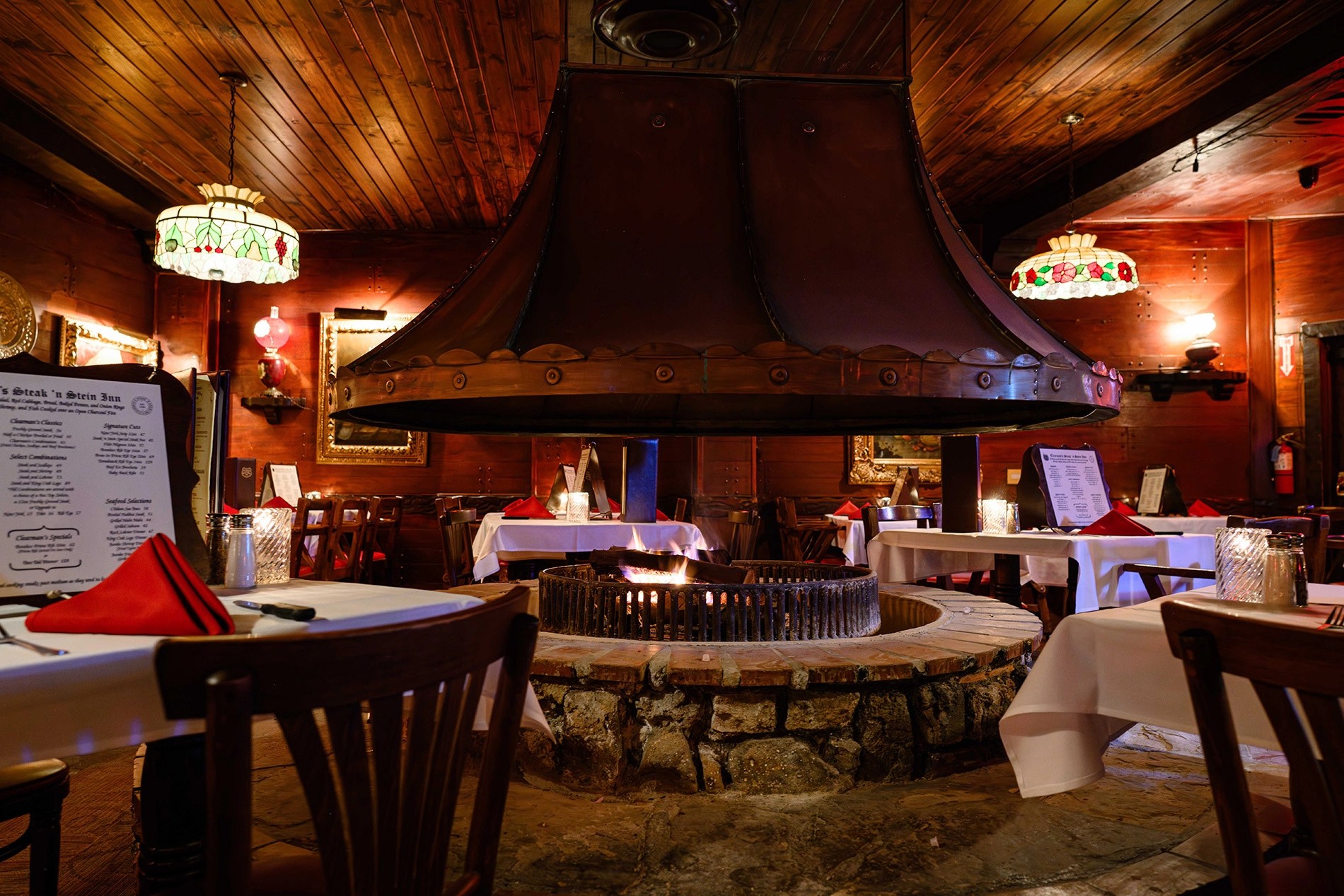 A cozy restaurant interior featuring a central circular fireplace with a metal hood, surrounded by wooden tables set with red napkins. Decorative lighting enhances the warm atmosphere.