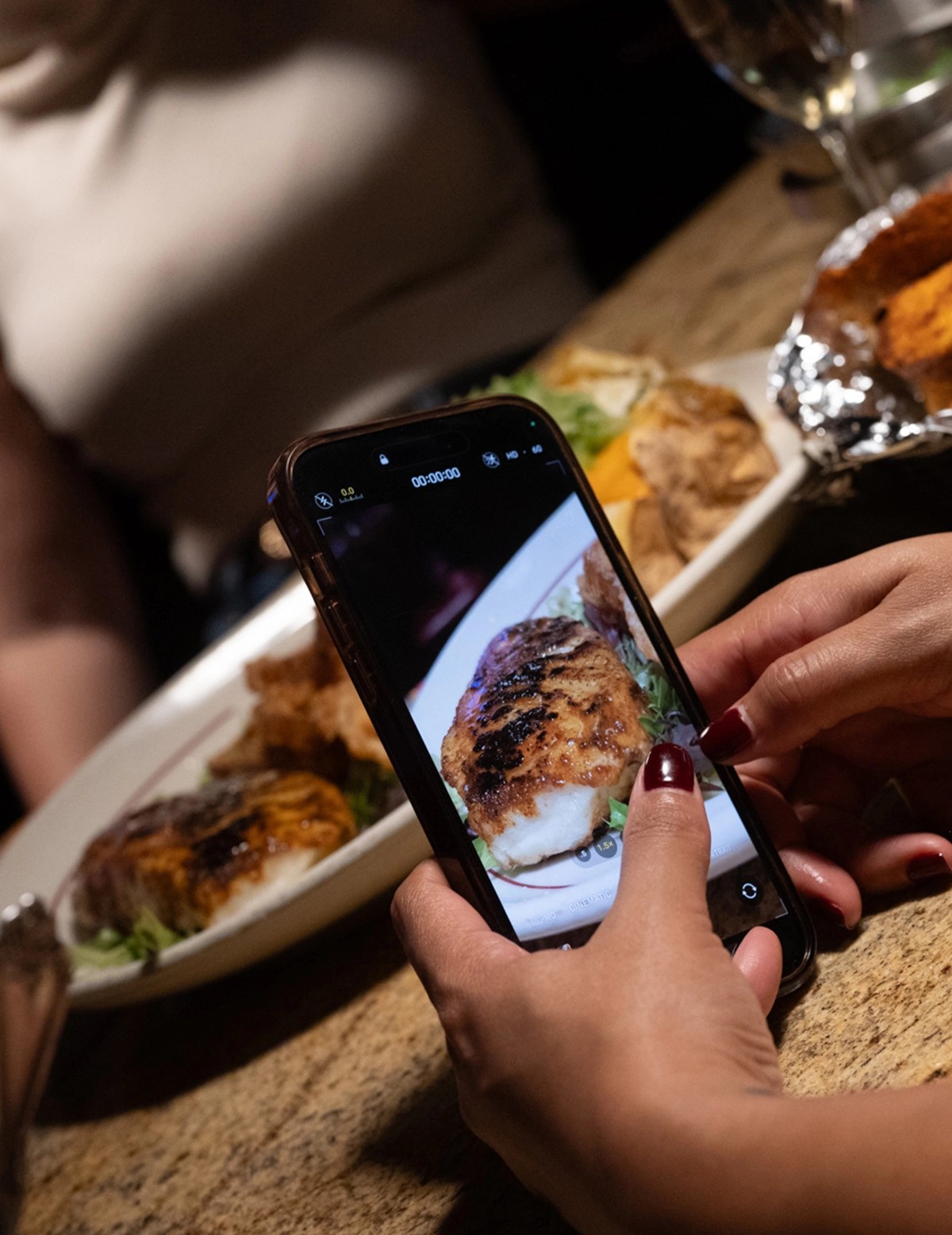 A person is taking a photo of a grilled fish dish on their phone, with a blurred background of food and a glass of white wine.