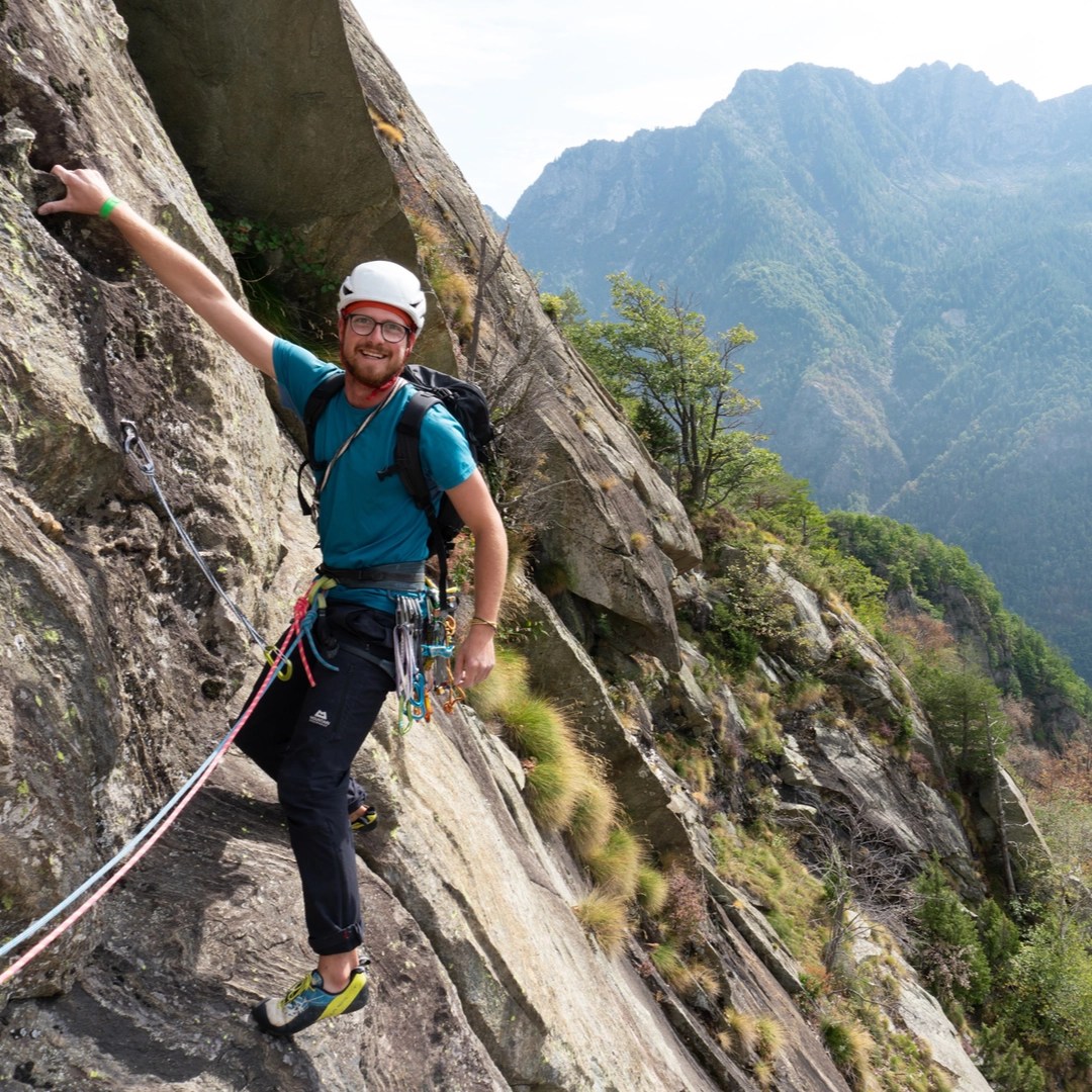 Andreas, a climbing guide and instructor, ascends a steep rock face in Italy. Wearing a teal shirt and white helmet, he is equipped with a harness full of climbing gear, against a backdrop of dense, green mountain forests.