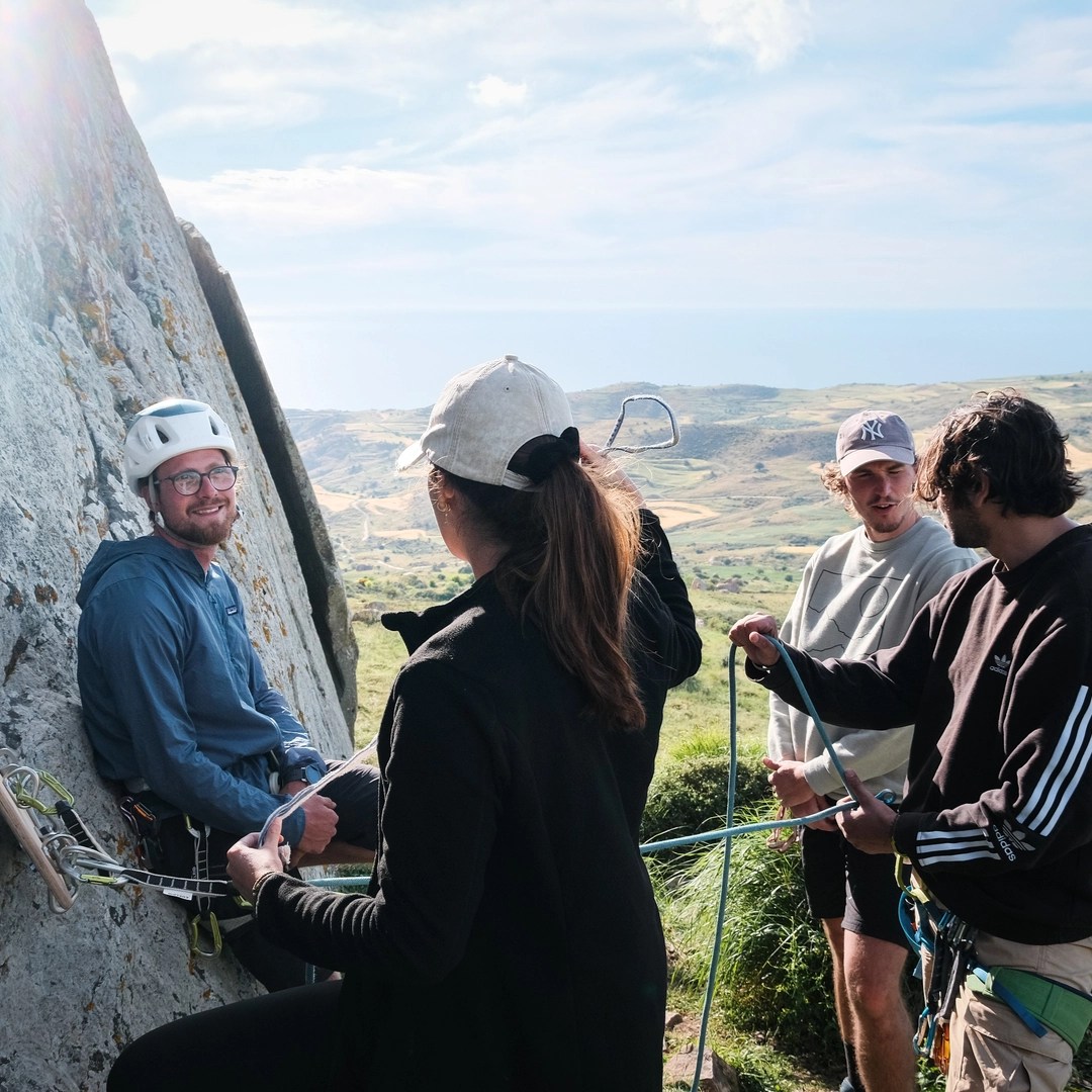 A group of young climbers is gathered at the base of a large rock face, with one climber, seated and wearing a helmet, harness, and glasses, smiling at the camera. The other three individuals, including a woman in a light-colored cap and a man in a black jacket, are engaged in a discussion, possibly receiving instructions. The background reveals a picturesque landscape with rolling hills and the sea in the distance under a partly cloudy sky, adding a serene and expansive feel to the scene.