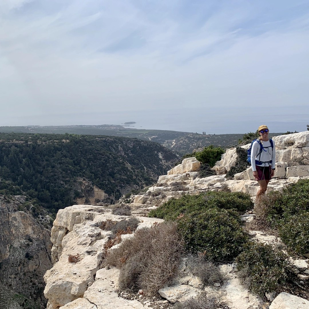 A person is standing on a rocky ledge in a rugged, coastal landscape, with expansive views of green hills, cliffs, and the distant sea. The individual is wearing hiking attire, including a hat, sunglasses, and a backpack, and seems to be enjoying the outdoor adventure. The sky is slightly overcast, casting a soft light over the terrain. The ledge they are standing on is surrounded by low shrubs and rocky outcrops, with the backdrop of a vast, undulating landscape leading down to the coastline. This image captures the essence of hiking in a remote and scenic area, likely a coastal trail with impressive views.
