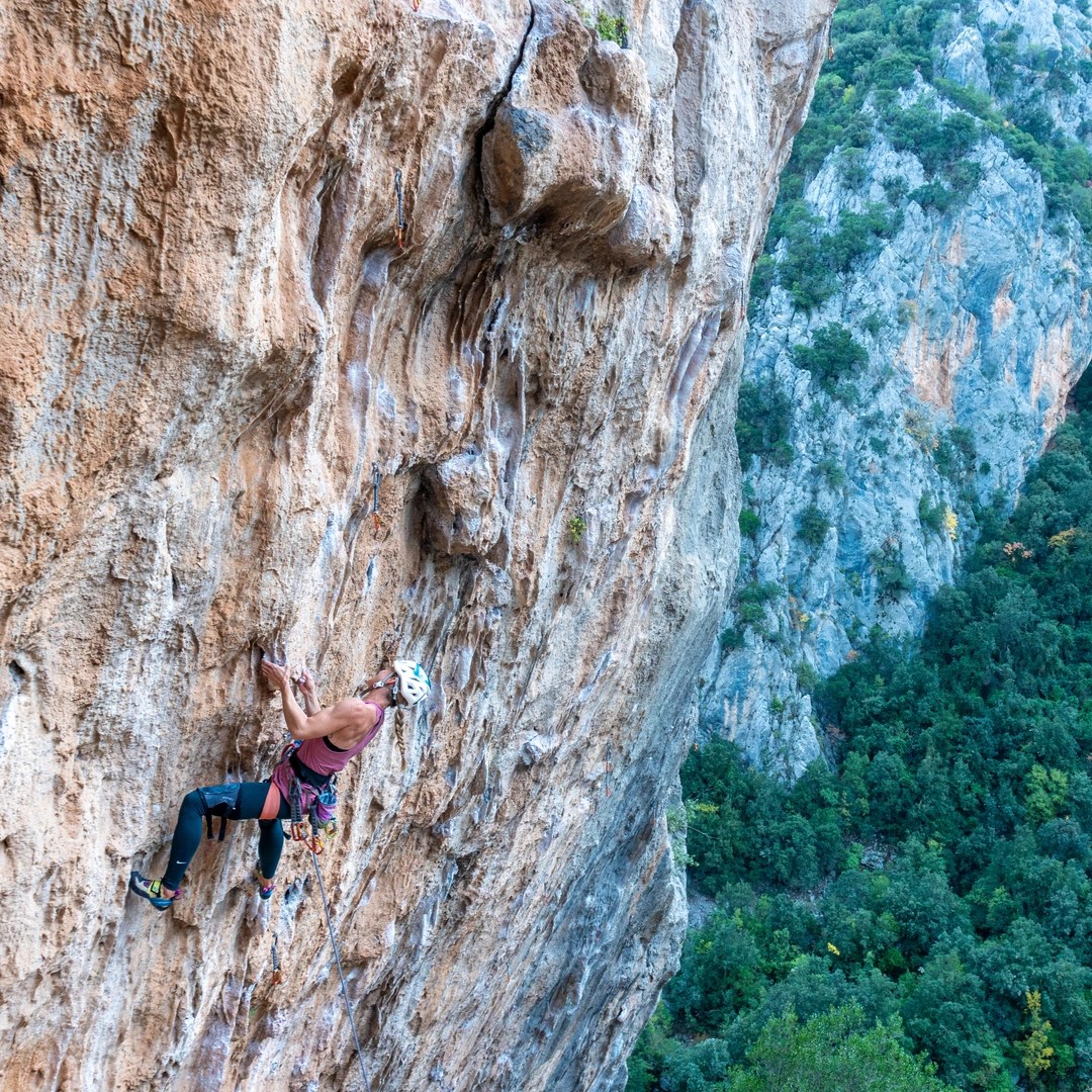 A female climber wearing a helmet, tank top, and knee pads is scaling a steep, overhanging rock face. The rock is orange-brown with visible chalk marks and quickdraws clipped to bolts. The background shows a steep, forested hillside, emphasizing the climber's height and the challenging nature of the climb. The scene likely takes place in a natural outdoor climbing area.