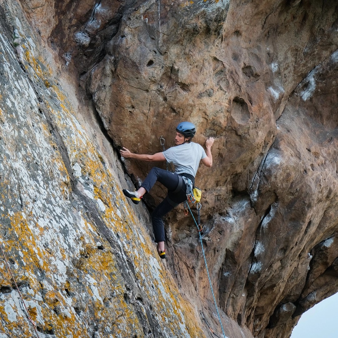 A climber, wearing a helmet and grey t-shirt, is captured mid-climb on a steep, textured rock face. The climber is carefully navigating a section with both hands and feet engaged, displaying a mix of concentration and determination. The rock is marked with lichen and features a series of small cracks and holds. The background shows the rugged and overhanging rock formation, highlighting the challenge of the climb. The climber is secured by a rope attached to a harness, indicating that this is a lead climbing scenario.