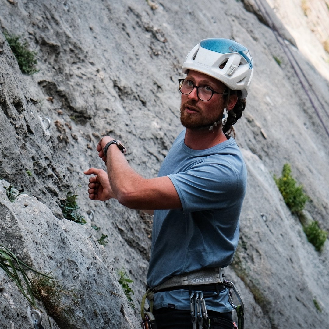 A person wearing a white helmet and blue t-shirt is pointing to something on a rock face while explaining climbing techniques, with climbing gear visible on their harness.
