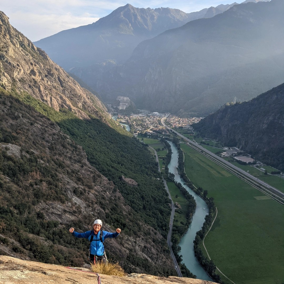 A person standing triumphantly on top of a cliff overlooking a valley with a winding river, surrounded by towering mountains.