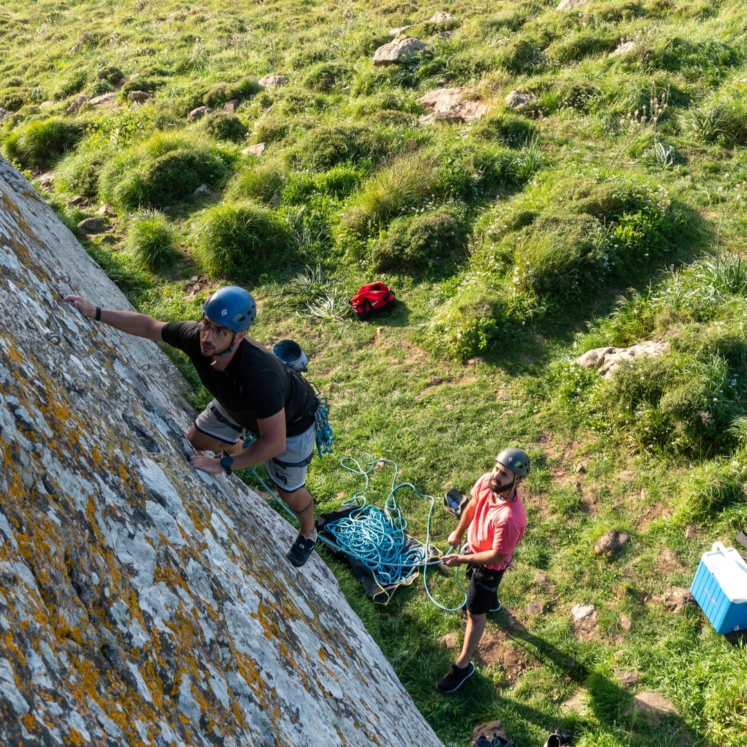 A climber is ascending a steep, lichen-covered rock face, wearing a blue helmet and black shirt. The climber is focused on placing their hand on a hold while preparing to clip the rope into a quickdraw attached to the rock. Below, another person, wearing a red shirt and helmet, stands on the grassy ground, belaying the climber and managing the rope. The lush green grass surrounding the base of the climb contrasts with the rugged texture of the rock face, creating a picturesque outdoor climbing scene. Climbing gear and ropes are visible on the ground beside the belayer.