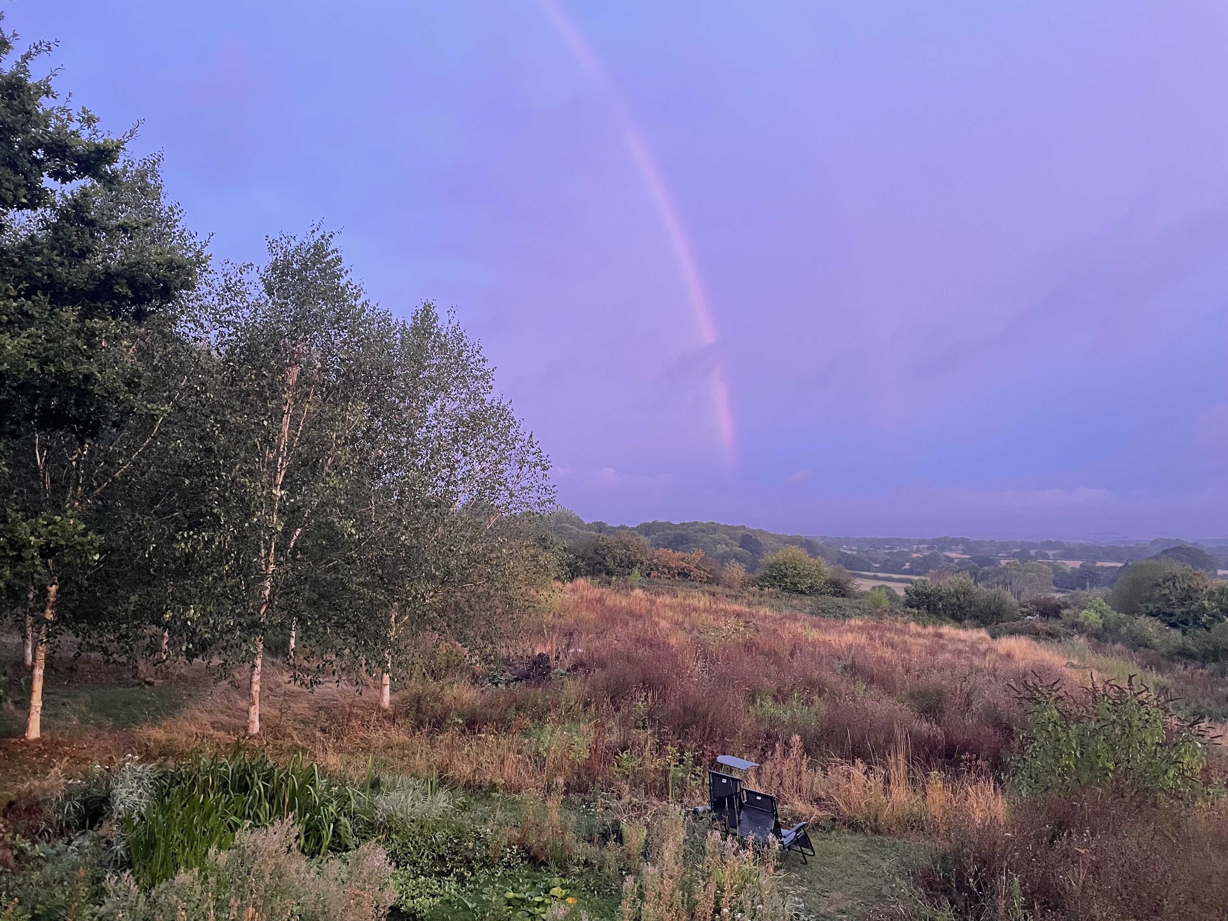 A faint rainbow arcs through a lavender-blue morning sky above a late-summer meadow, with trees, fields and distant woodland beneath.