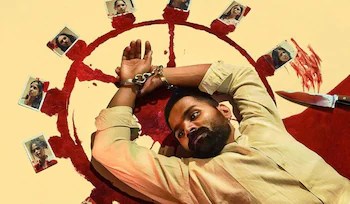 High-angle shot of the character Stephen lying on the floor with his hands handcuffed above his head, surrounded by a red painted circle containing photographs of female victims and a knife.