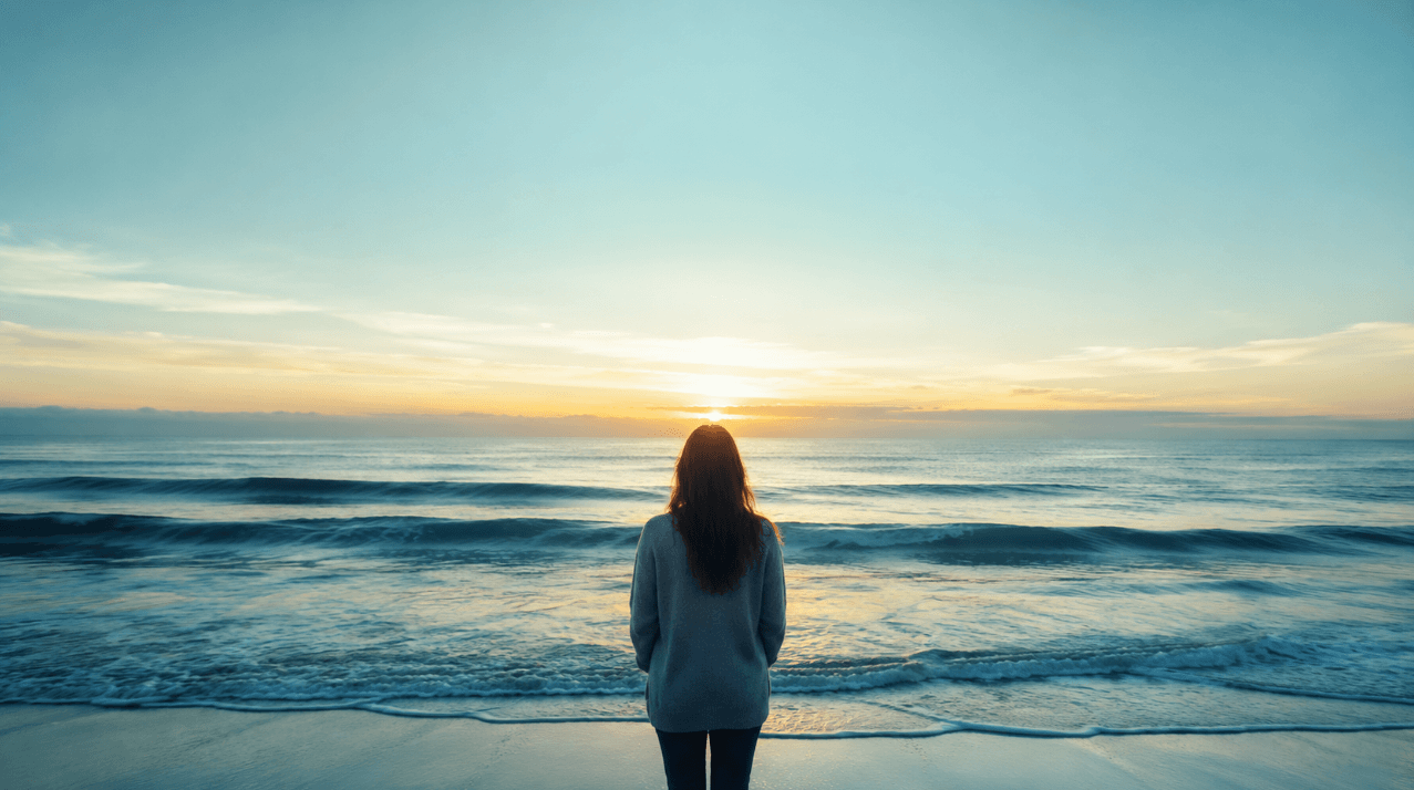 A person standing peacefully at the shoreline at dawn, calm teal waves ahead, choosing support for grief and healing.