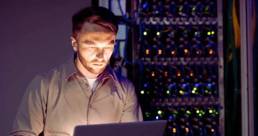 IT manager looking at computer standing by rack of servers - The Cogle Group