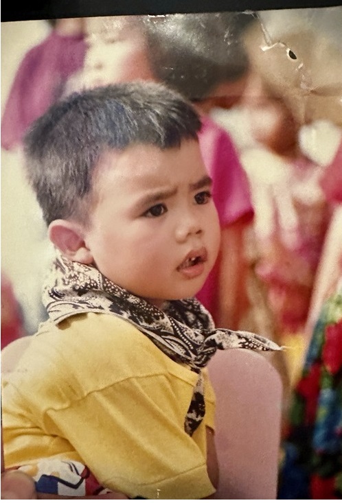 A childhood photo of Khris Baizen. He is wearing a bright yellow shirt with a patterned scarf tied around his neck. He appears to be sitting down with other children blurred in the background. His expression is serious and inquisitive, with furrowed brows and slightly parted lips.