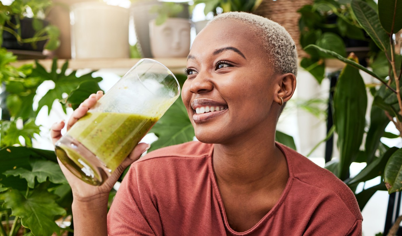 A young woman sits in a room surrounded by plants, sips a smoothie from a glass