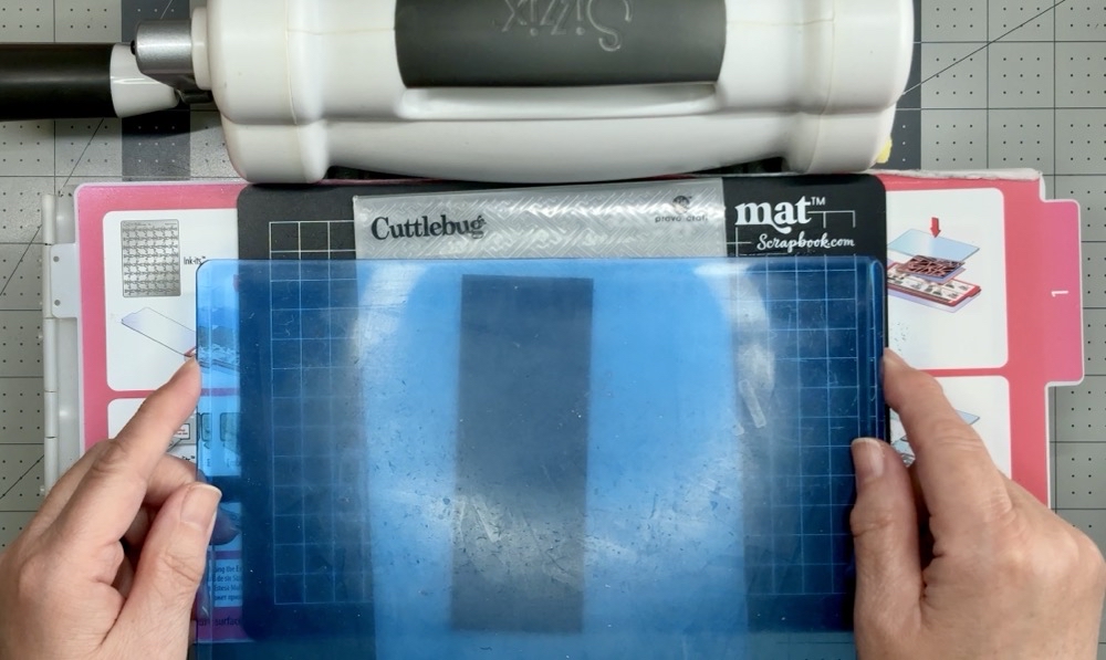 Photo showing the sandwich needed to emboss the Cuttlebug embossing folder with the Sizzix die cutting machine consisting of the base plate, shim #1, scrapbook.com magic mat, embossing folder and card stock, and a final top cutting plate.