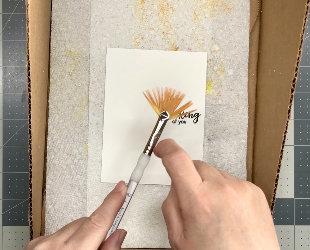 Photo showing how I hold the brush in one hand and tap it lightly with my finger to add the subtle splatter spots to the card panel for todayโs One Card Design, Endless Creative Looks projects.
